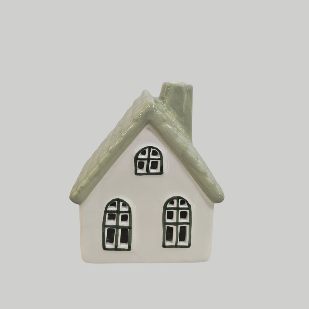 A small ceramic house-shaped container or candle holder with a white body, textured sage green roof, and black window outlines, shown against a solid light gray background.