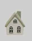 A small ceramic house-shaped container or candle holder with a white body, textured sage green roof, and black window outlines, shown against a solid light gray background.