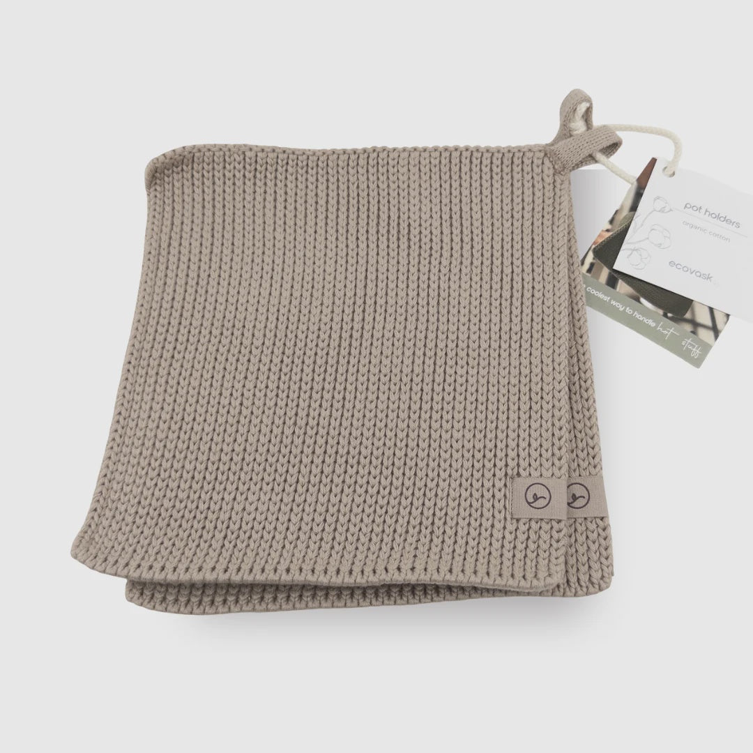 A folded chunky knit woolen scarf in a light taupe-beige color, shown with its paper hangtag on a clean white background.