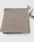 A folded chunky knit woolen scarf in a light taupe-beige color, shown with its paper hangtag on a clean white background.