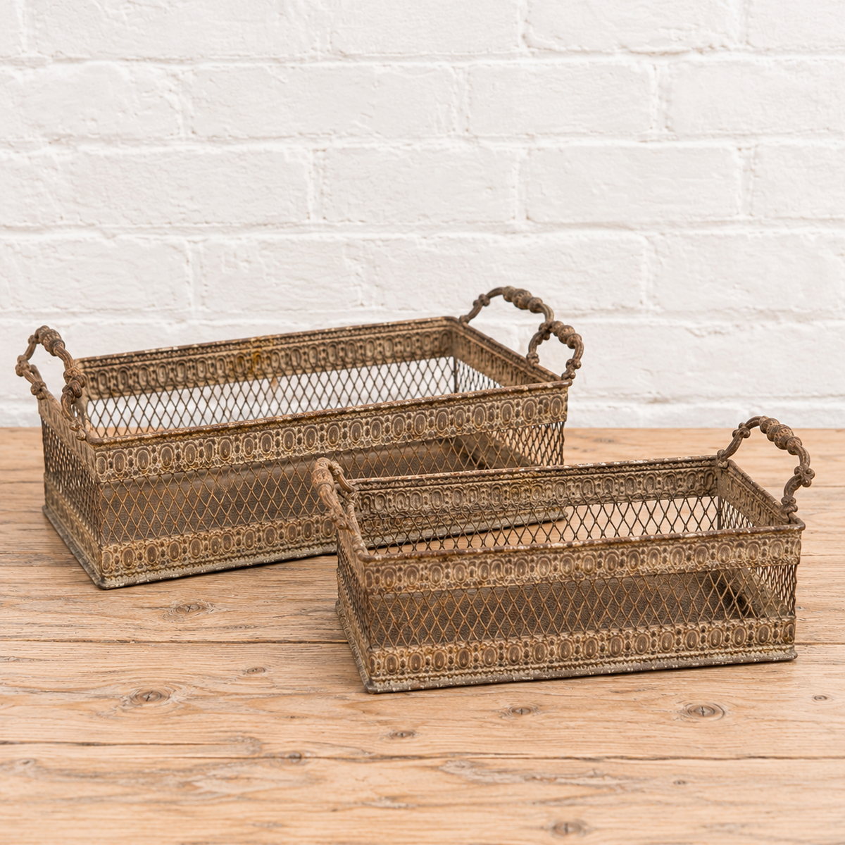 Two rectangular metal wire storage baskets with decorative patterns and rope handles, displayed on a wooden surface against a white brick wall.