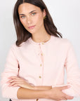 Woman wearing a light pink cardigan with gold buttons on a plain background