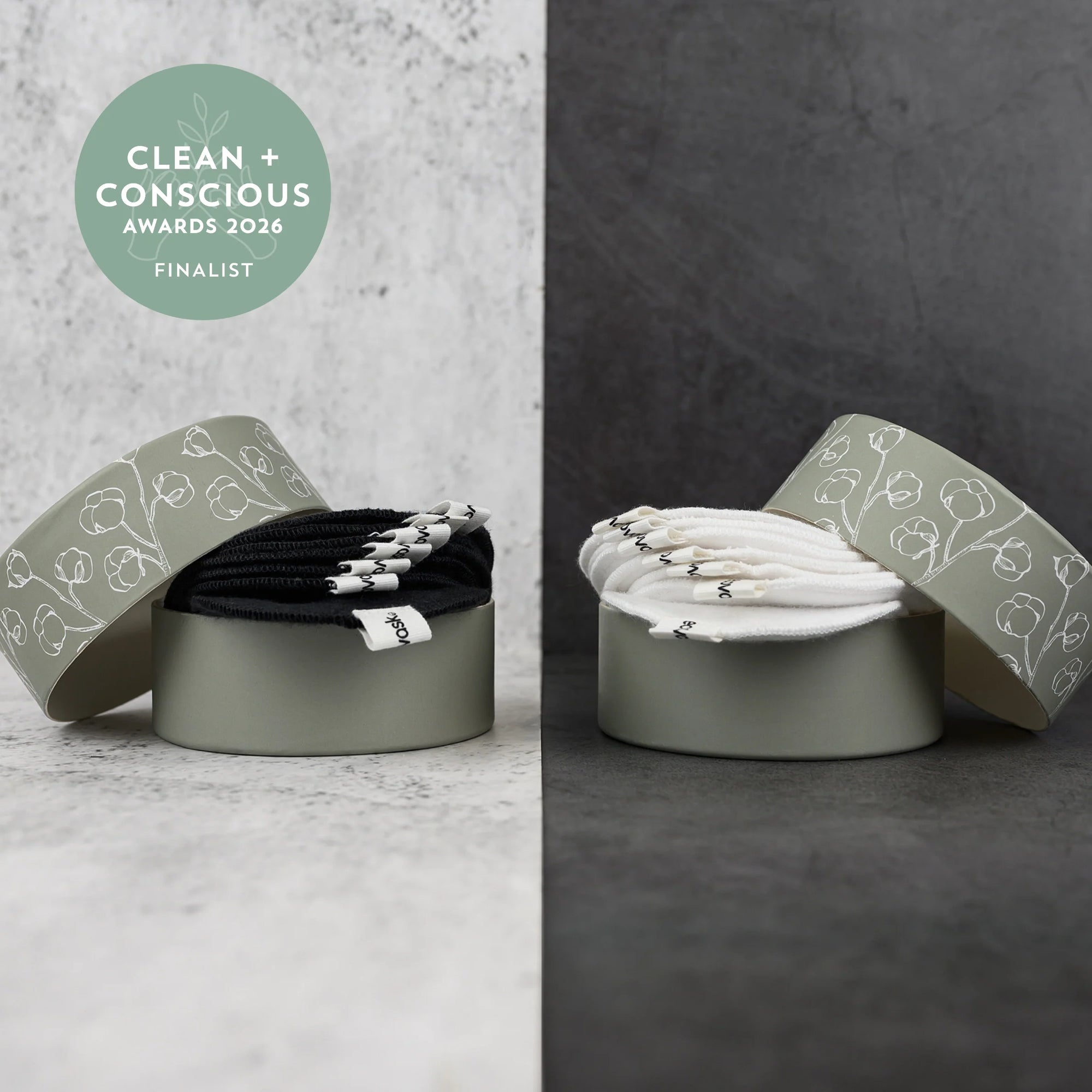 Two round metal gift boxes with patterned lids, each containing a bundle of elastic hair ties. The left box shows black hair ties with silver metal ends, and the right box shows white hair ties with black ends.