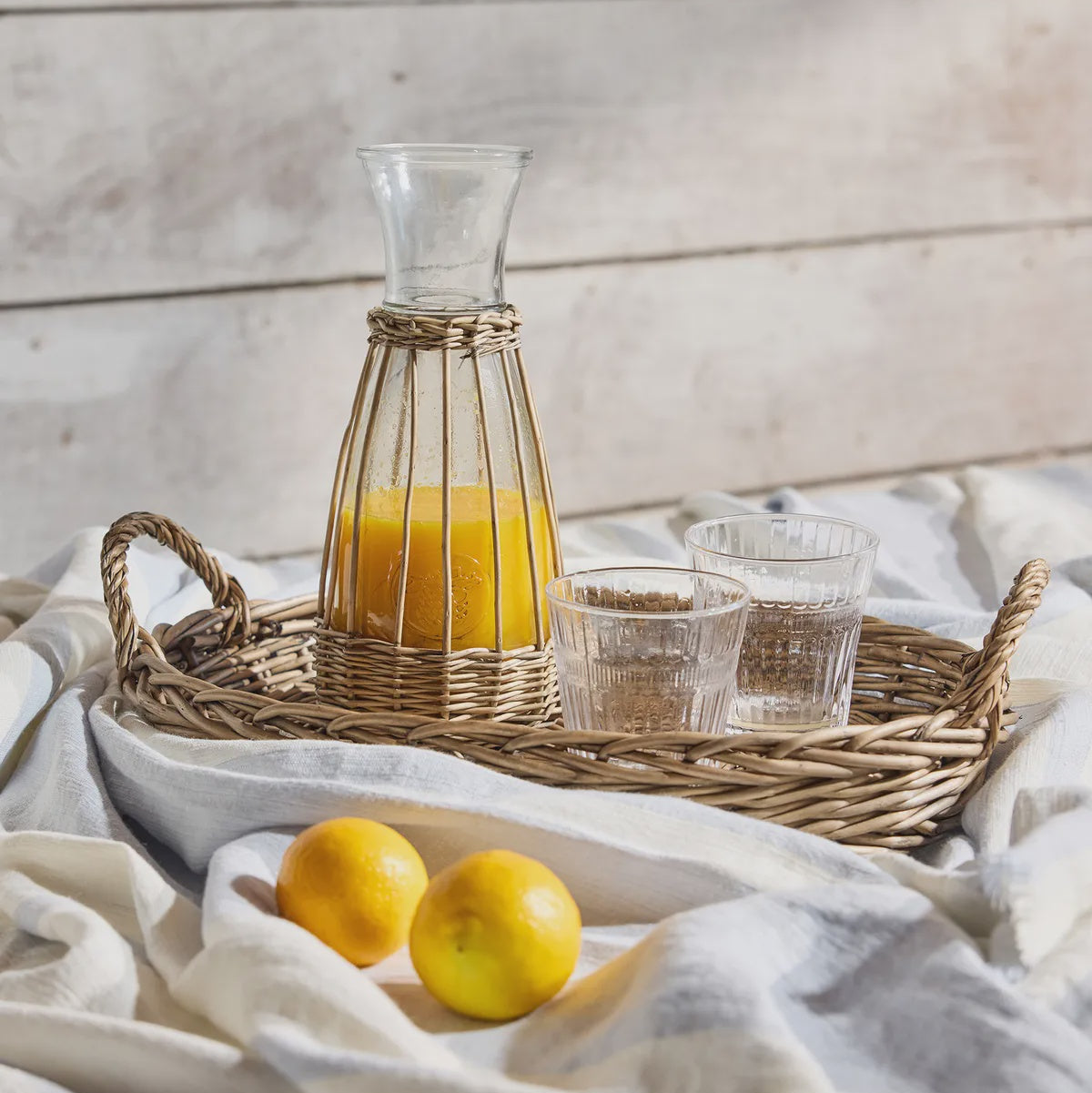 Carafe of orange juice on a woven tray with glasses and lemons on a white cloth against a wooden background