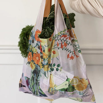 Reusable shopping bag with floral and fruit design filled with groceries, held by a person.