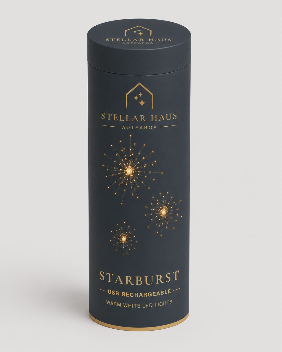 A tall cylindrical product box for Stellar Haus Agtearoa Starburst USB rechargeable warm white LED lights, featuring a dark charcoal gray body with gold foil starburst graphics and text, set against a light gray background.