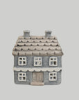A small ceramic house-shaped container or figurine with a gray-blue body, brown roof, and white window details, centered on a plain light gray background.