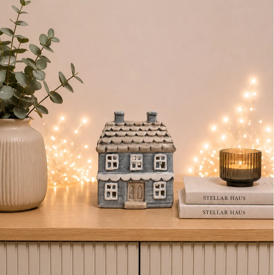 A small gray ceramic house figurine sits on a light wood cabinet, styled with a beige ribbed vase holding eucalyptus leaves on the left and a stack of books with a lit candle on the right against a warm beige wall with string lights.