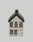 A small decorative house-shaped ceramic candle holder or ornament with a black roof, black window frames, and a white body with subtle gray horizontal striping, centered on a light gray background.