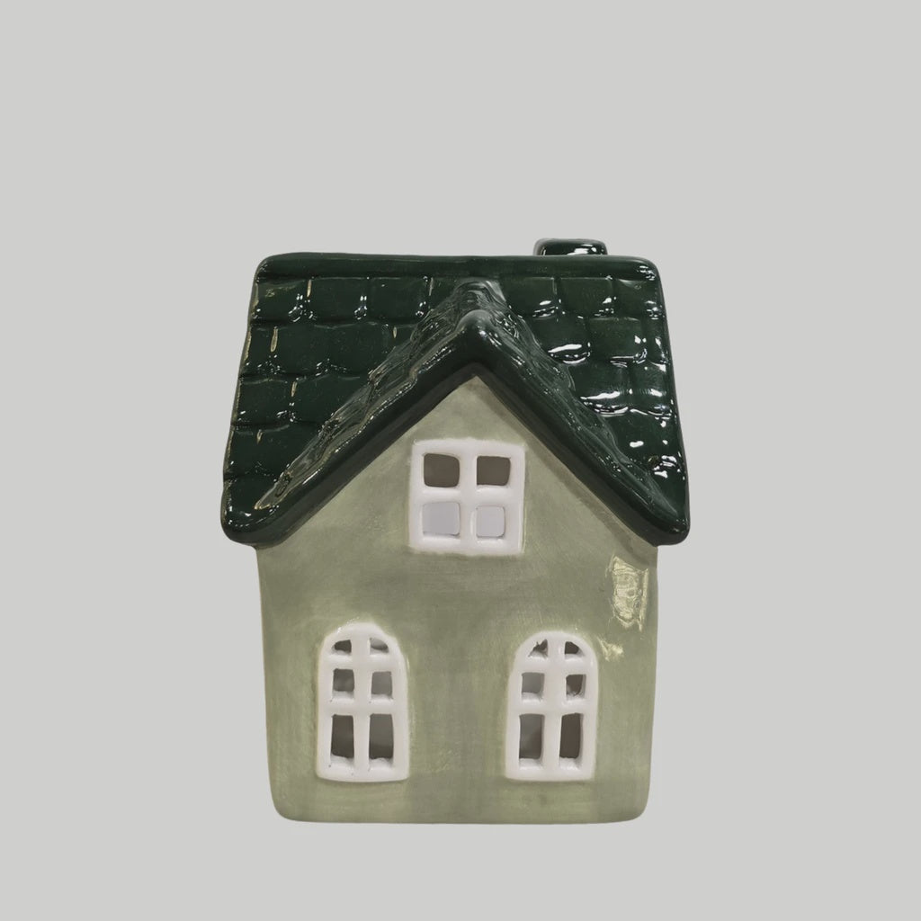 A small decorative house-shaped ceramic container or ornament with a dark green textured roof and a light sage green body, shown against a plain light gray background.