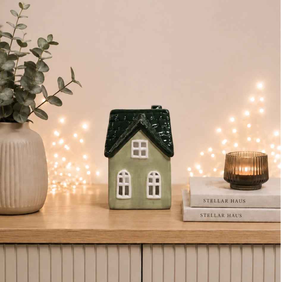 A small ceramic house-shaped candle holder in sage green with a dark green textured roof and white window details, displayed on a light wood cabinet next to a potted eucalyptus plant and a stack of Stellar Haus books with a lit candle.