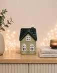 A small ceramic house-shaped candle holder in sage green with a dark green textured roof and white window details, displayed on a light wood cabinet next to a potted eucalyptus plant and a stack of Stellar Haus books with a lit candle.