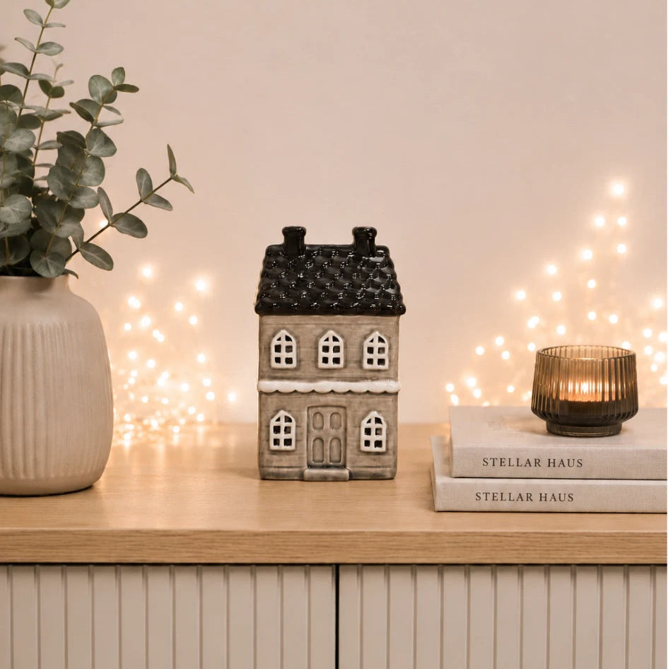A small decorative house-shaped ceramic candle holder with a dark textured roof and white window details sits on a light wood cabinet. The scene is styled with a beige vase of greenery on the left, a lit candle in a ribbed glass holder on books to the right, and warm fairy lights glowing in the background against a soft beige wall.