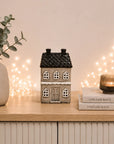 A small decorative house-shaped ceramic candle holder with a dark textured roof and white window details sits on a light wood cabinet. The scene is styled with a beige vase of greenery on the left, a lit candle in a ribbed glass holder on books to the right, and warm fairy lights glowing in the background against a soft beige wall.