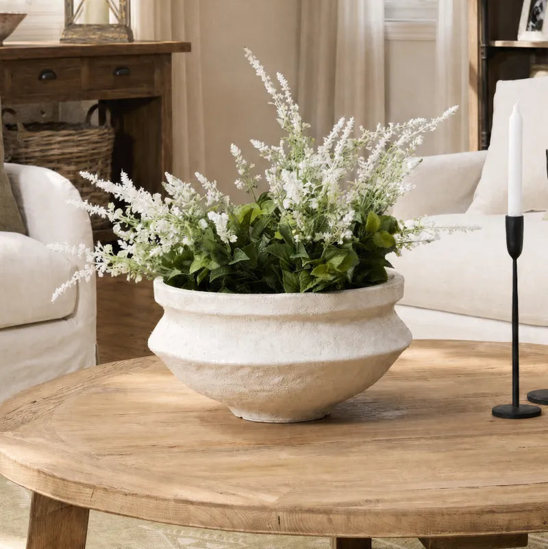 A rustic white ceramic planter filled with green foliage and white flowers, displayed on a round wooden coffee table in a cozy living room setting.