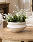 A rustic white ceramic planter filled with green foliage and white flowers, displayed on a round wooden coffee table in a cozy living room setting.