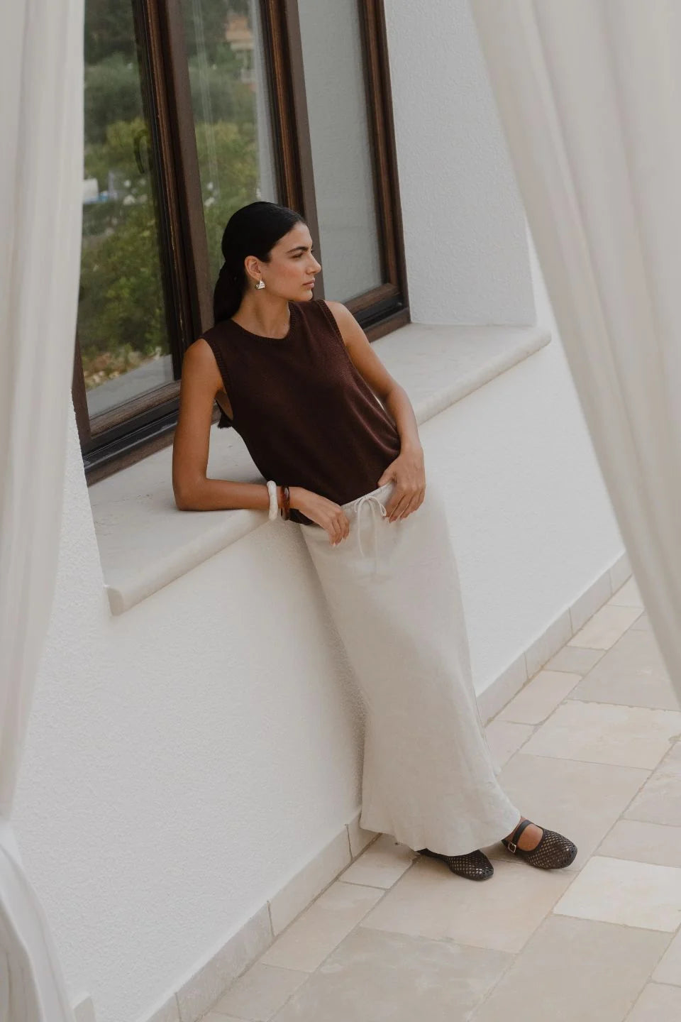 A woman wearing a sleeveless dark brown top and a long cream skirt leans against a white window ledge in a bright, modern interior.
