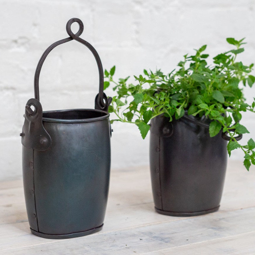 Two dark metal hanging planters with arched handles, one empty and one filled with a leafy green plant, set on a light wooden surface against a white brick wall.