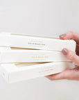 Hand holding a stack of white boxes with gold text on a light gray background