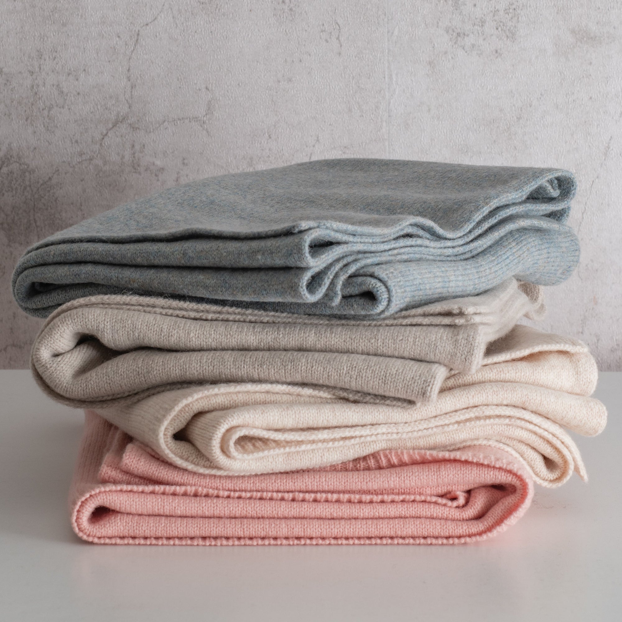 Stack of folded blankets in blue, beige, and pink on a light surface with a neutral background