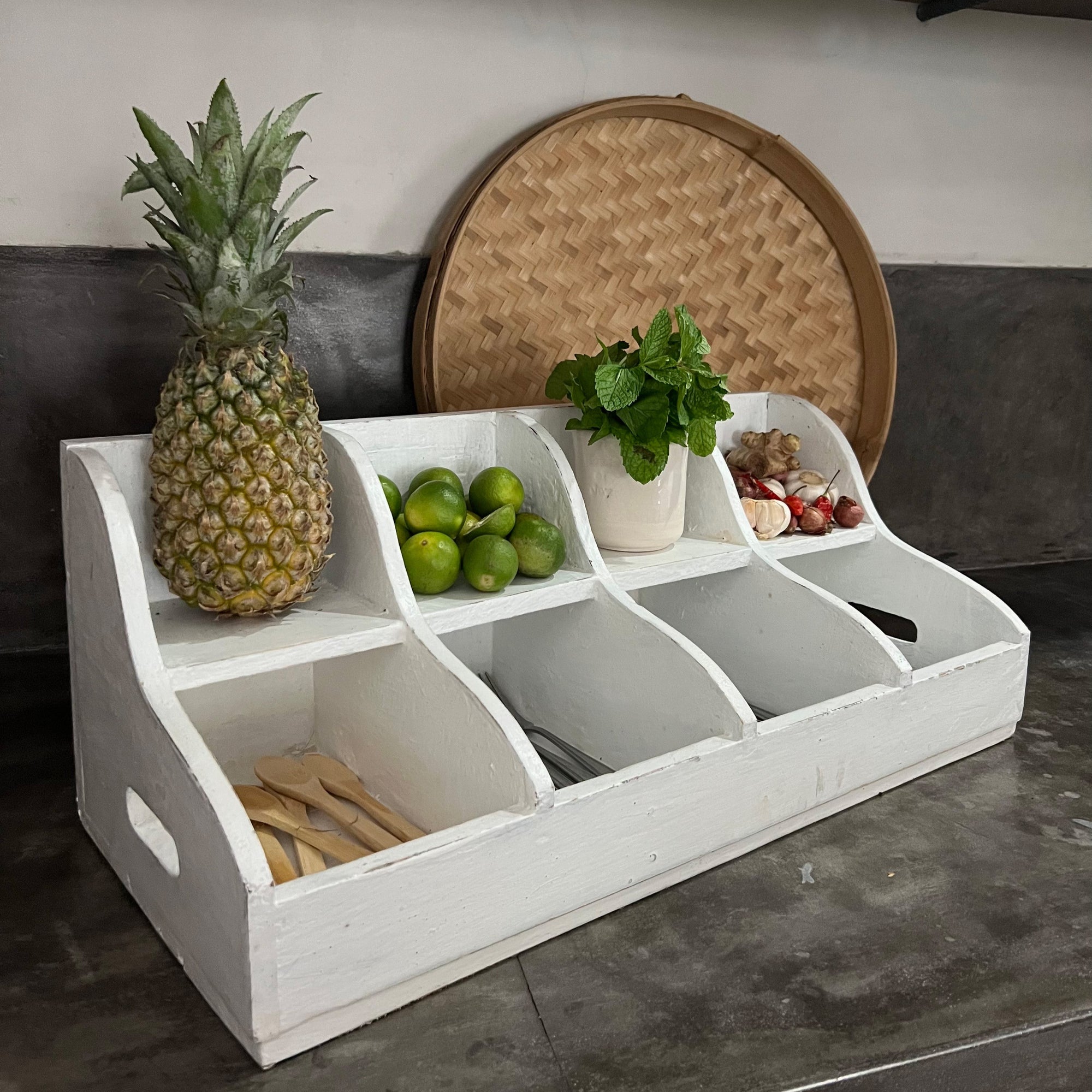 White wooden fruit basket with compartments containing a pineapple, limes, and other items on a kitchen counter.