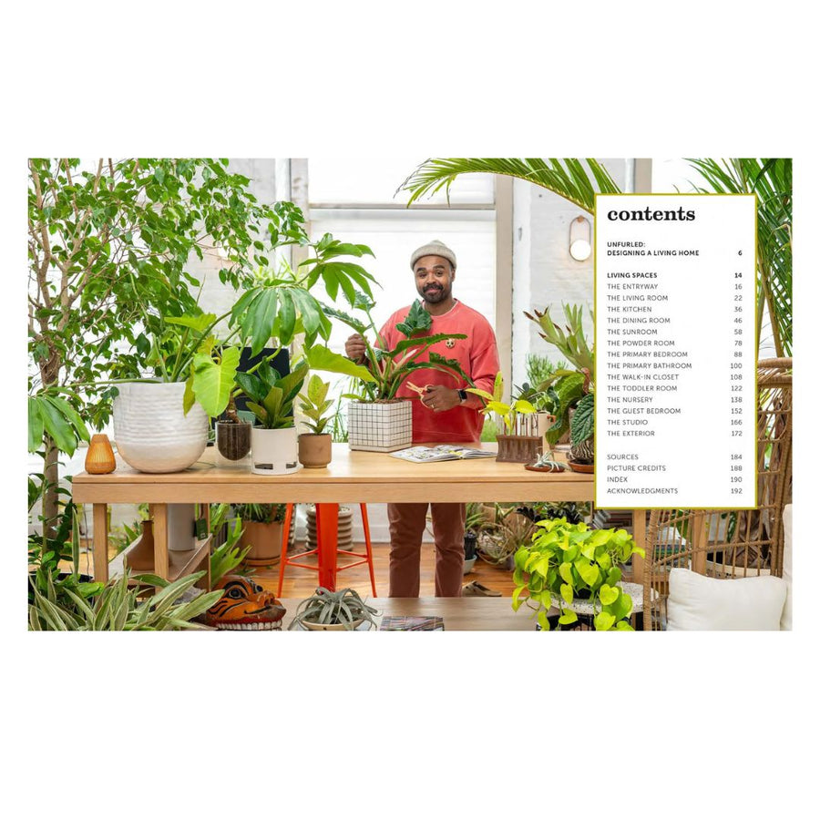 Person in a room filled with plants, holding a plant, with a table and book visible.