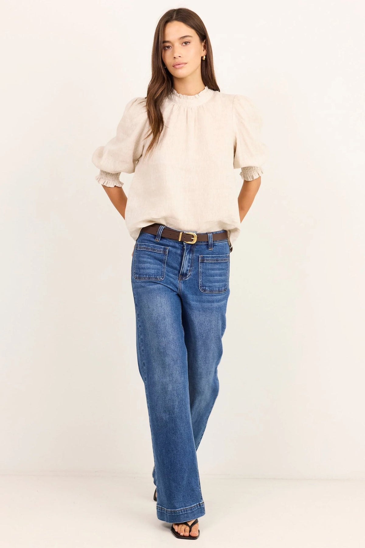 A woman modeling a cream puff-sleeve blouse and blue wide-leg jeans with a brown belt, standing against a plain off-white studio background.