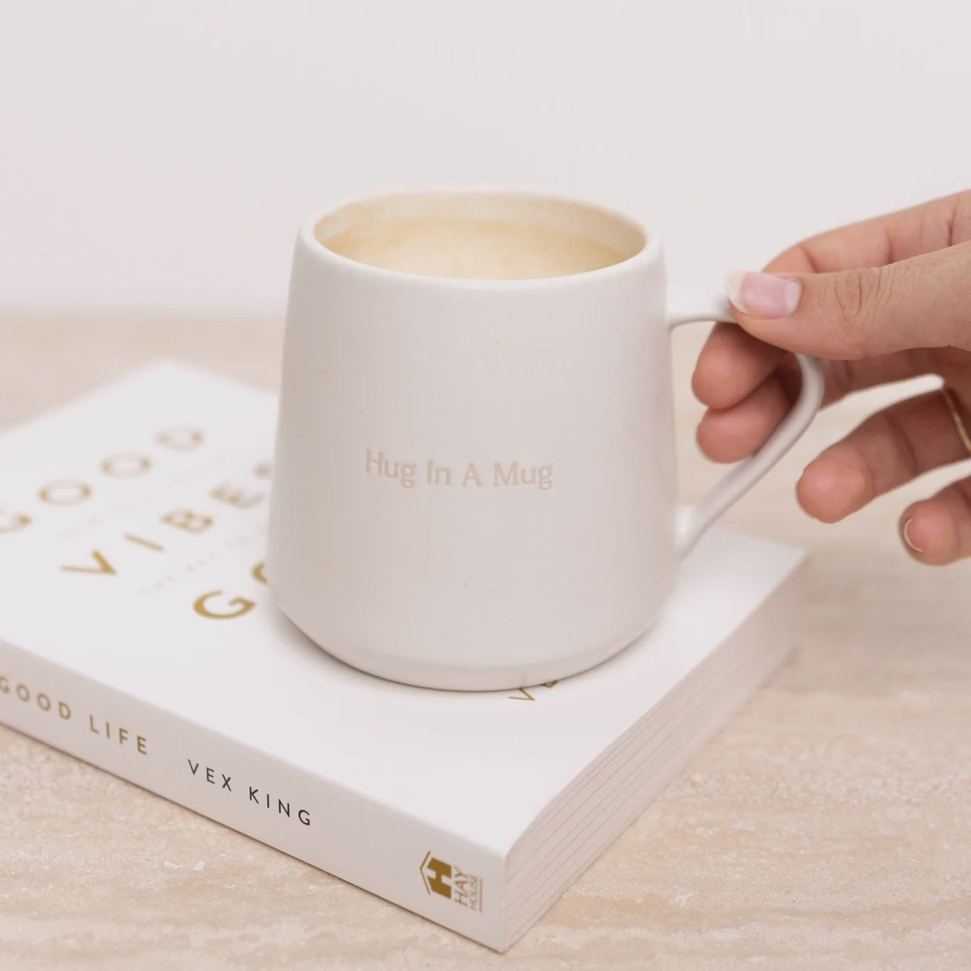 White mug with 'Hug in a Mug' text held over an open book on a light surface.
