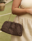 Woman holding a woven handbag in an outdoor setting with columns and plants.