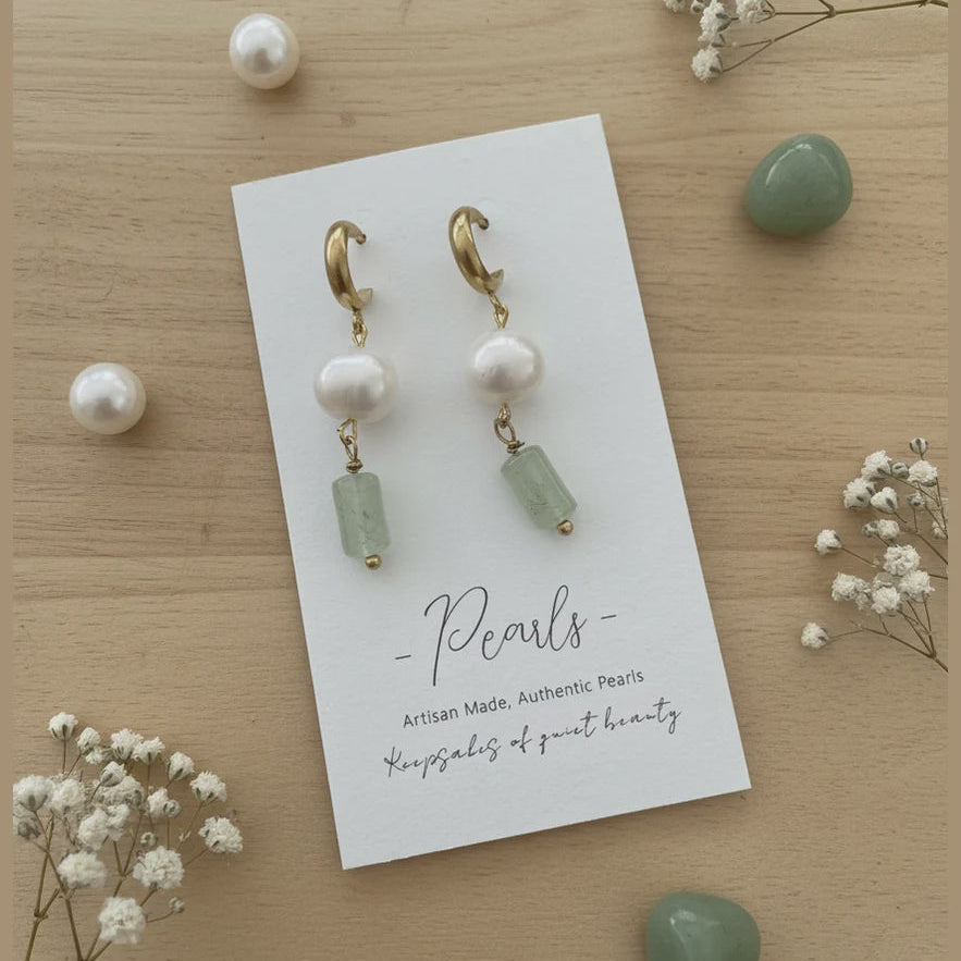 A pair of drop earrings displayed on a white card labeled 'Pearls' on a light wood surface, surrounded by decorative pearls, green stones, and small white flowers. The earrings have gold-tone hoops, white pearl beads, and light green rectangular beads.