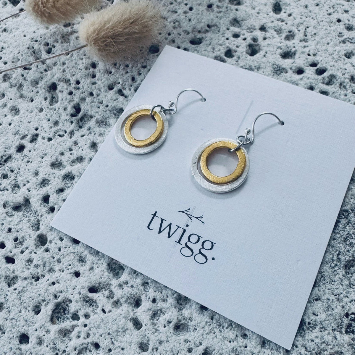 A pair of silver and gold hoop drop earrings displayed on a white Twigg brand card, set against a textured gray stone background with dried bunny tail grass.