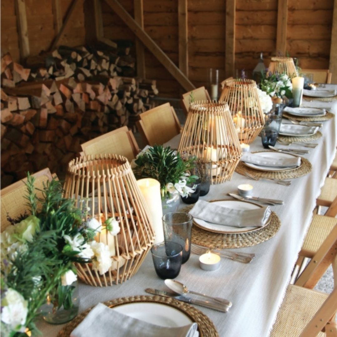 Dining table set with wicker lanterns, candles, and greenery in a rustic setting.