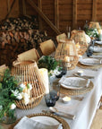 Dining table set with wicker lanterns, candles, and greenery in a rustic setting.