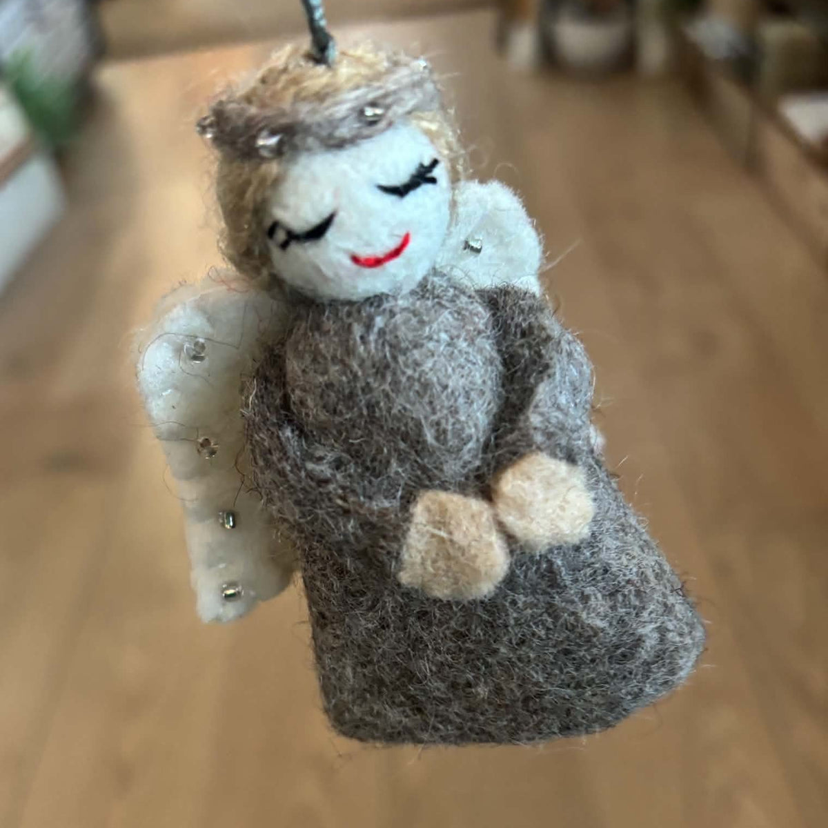 A handmade felt angel ornament with a gray dress, white wings, and a gold halo, hanging against a blurred wooden floor background.
