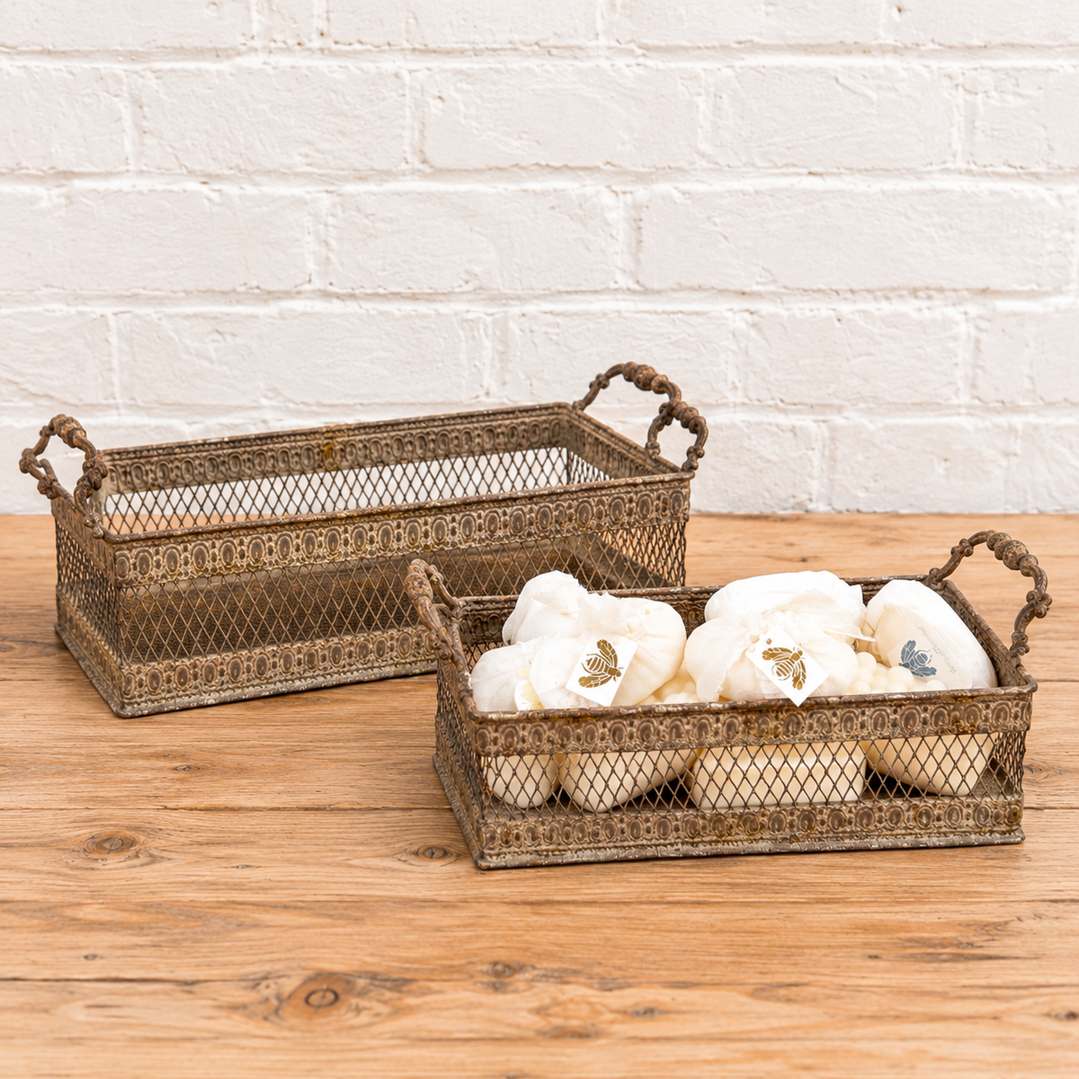 Two rectangular metal wire baskets with rope handles sit on a wooden surface against a white brick wall. The front basket holds several white decorative pillows or cushions.