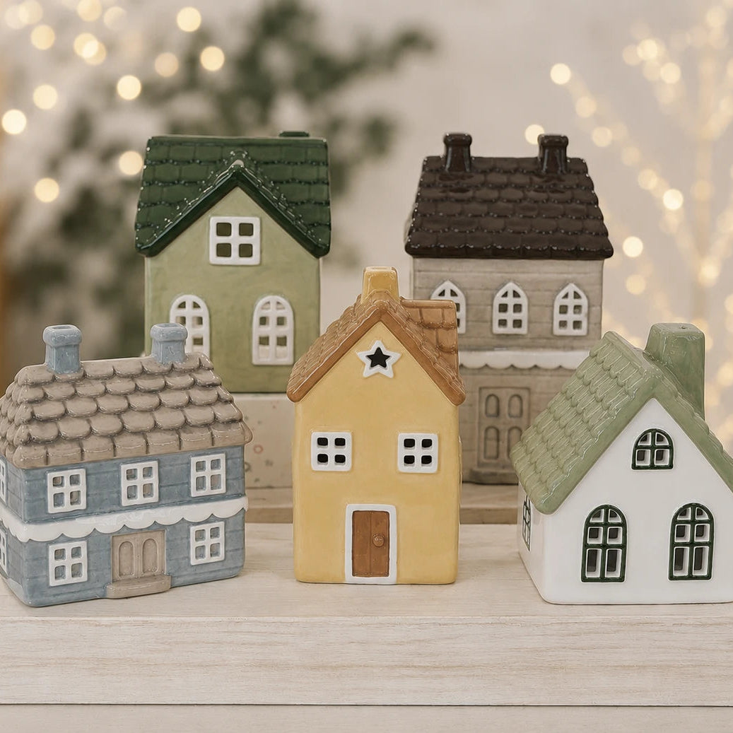 Five miniature ceramic house figurines in assorted colors and styles, arranged on a light-colored wooden block against a softly blurred background with warm bokeh lights.