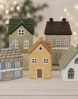 Five miniature ceramic house figurines in assorted colors and styles, arranged on a light-colored wooden block against a softly blurred background with warm bokeh lights.