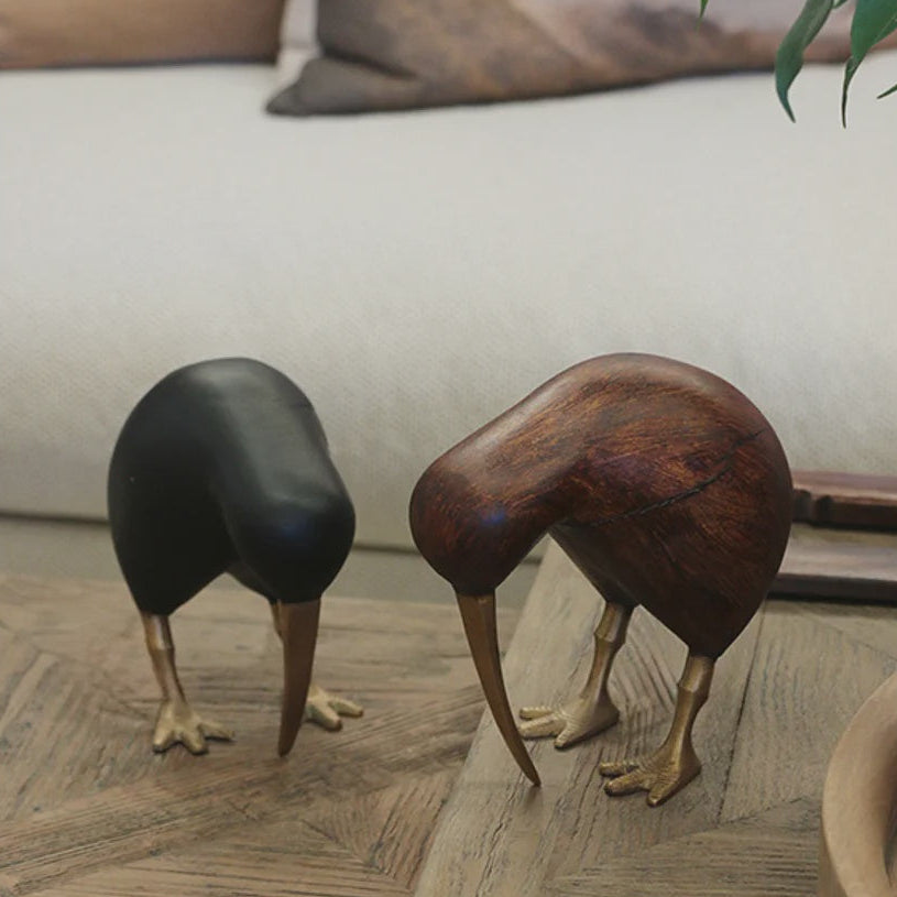 Two kiwi bird figurines on a wooden floor in a living room setting. One figurine is made of black wood, and the other is made of polished brown wood, both with gold-colored metal legs and beaks.